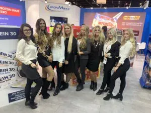 Acht Frauen posieren gemeinsam an einem Messestand mit Werbebannern und Displays im Hintergrund.