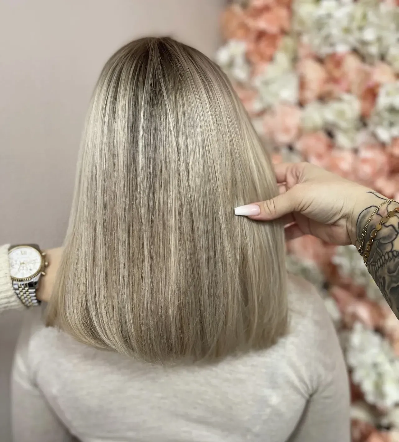 Eine Hand hält glattes, blondes, schulterlanges Haar vor einem unscharfen, floralen Hintergrund.