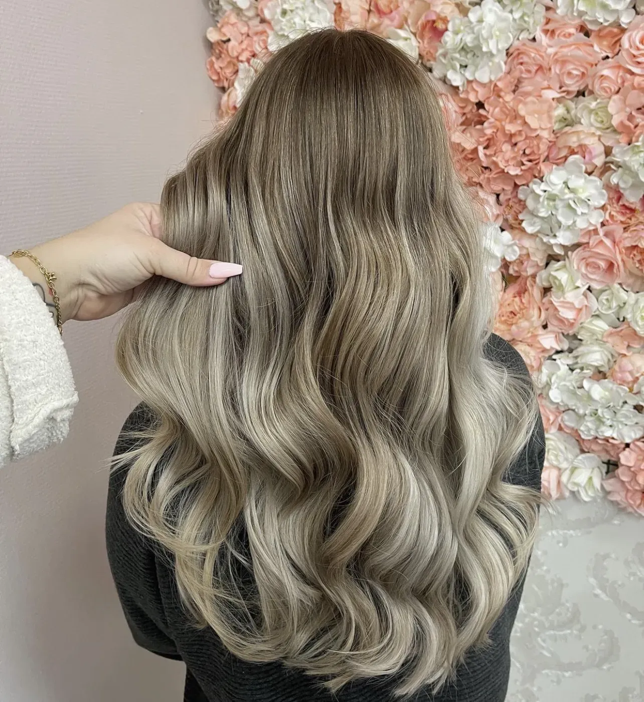 Eine Person mit langem, welligem, aschblondem Haar steht vor einer rosa-weißen Blumenwand.