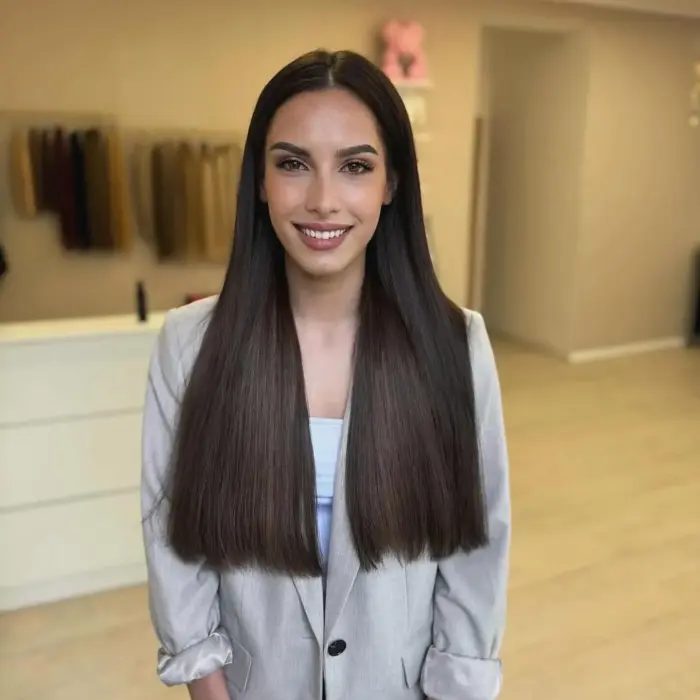 Eine Frau mit langem, glattem braunem Haar lächelt und trägt einen hellen Blazer in einem modernen, sanft beleuchteten Raum.