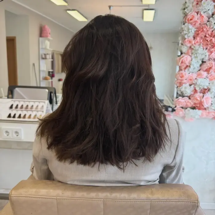 Eine Frau mit mittellangem, welligem, dunklem Haar sitzt in einem Friseurstuhl und blickt von der Kamera weg.