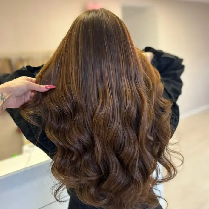 Frau mit langem, welligem, glänzendem braunem Haar, von hinten gesehen, in einer hellen Innenumgebung.