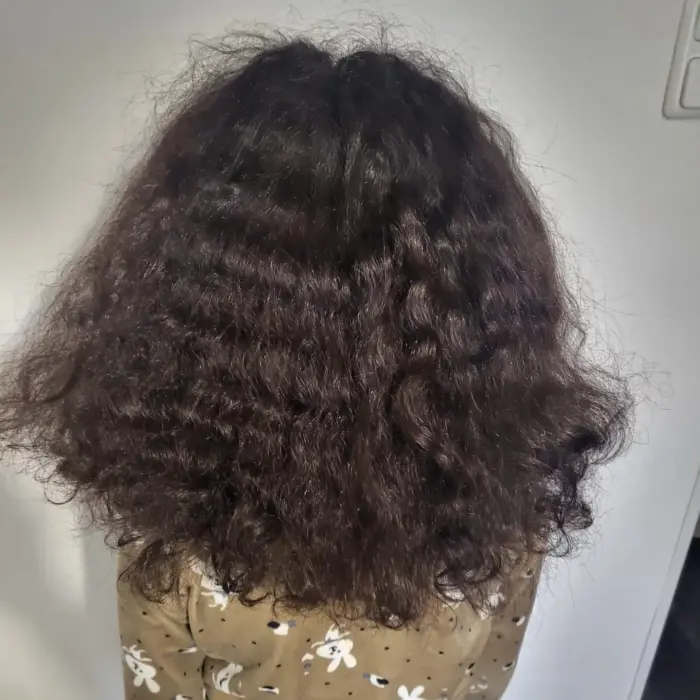 Ein Kind mit dichtem, lockigem braunem Haar steht vor einer weißen Wand und trägt ein braunes Hemd mit weißem Tiermuster.